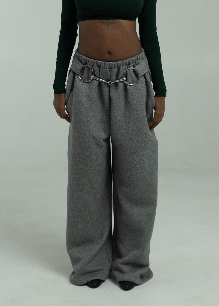 Person wearing a green long-sleeve top and gray wide-leg pants with horse bit on a plain background